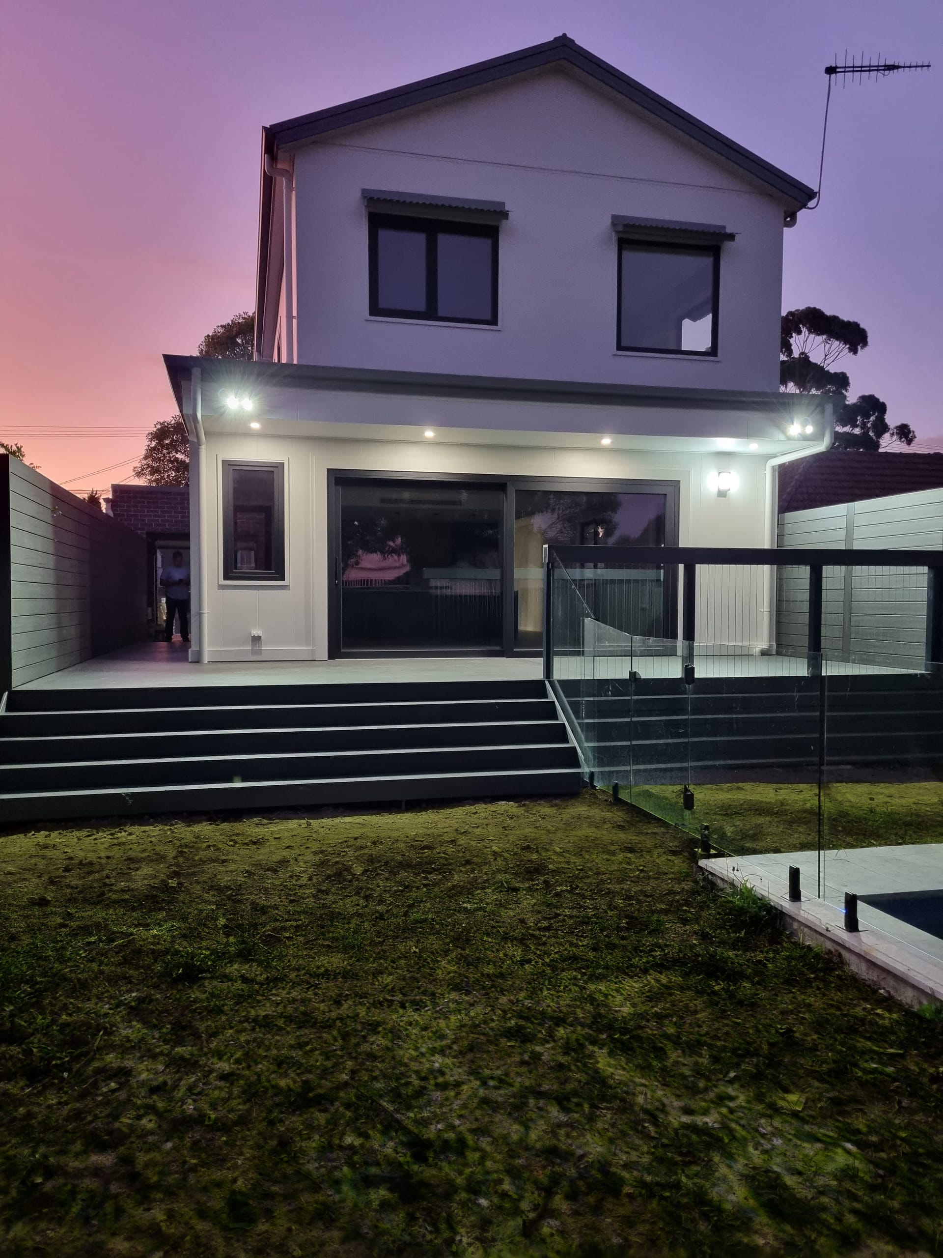 Rear facade at dusk, a white two-storey home with a covered alfresco, glass pool fencing and downlit eaves