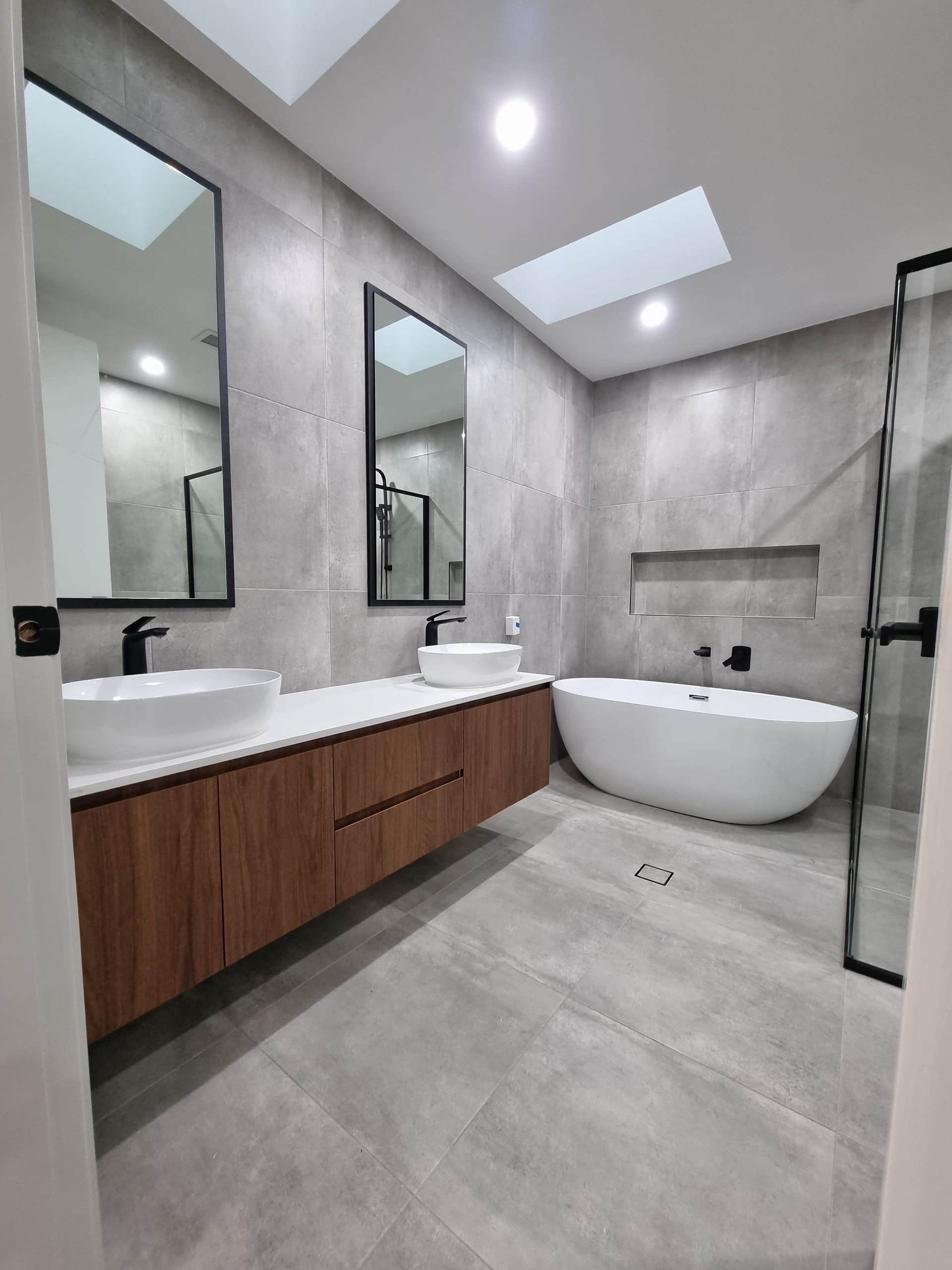 Main bathroom in large-format grey tiles with a double timber vanity, vessel basins, freestanding tub and a skylight