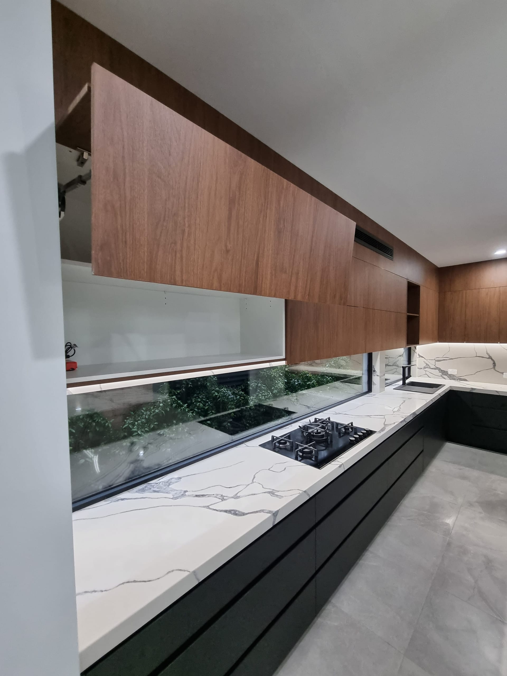 Galley kitchen run with a stone benchtop, gas cooktop, window splashback to greenery and timber overhead cabinetry