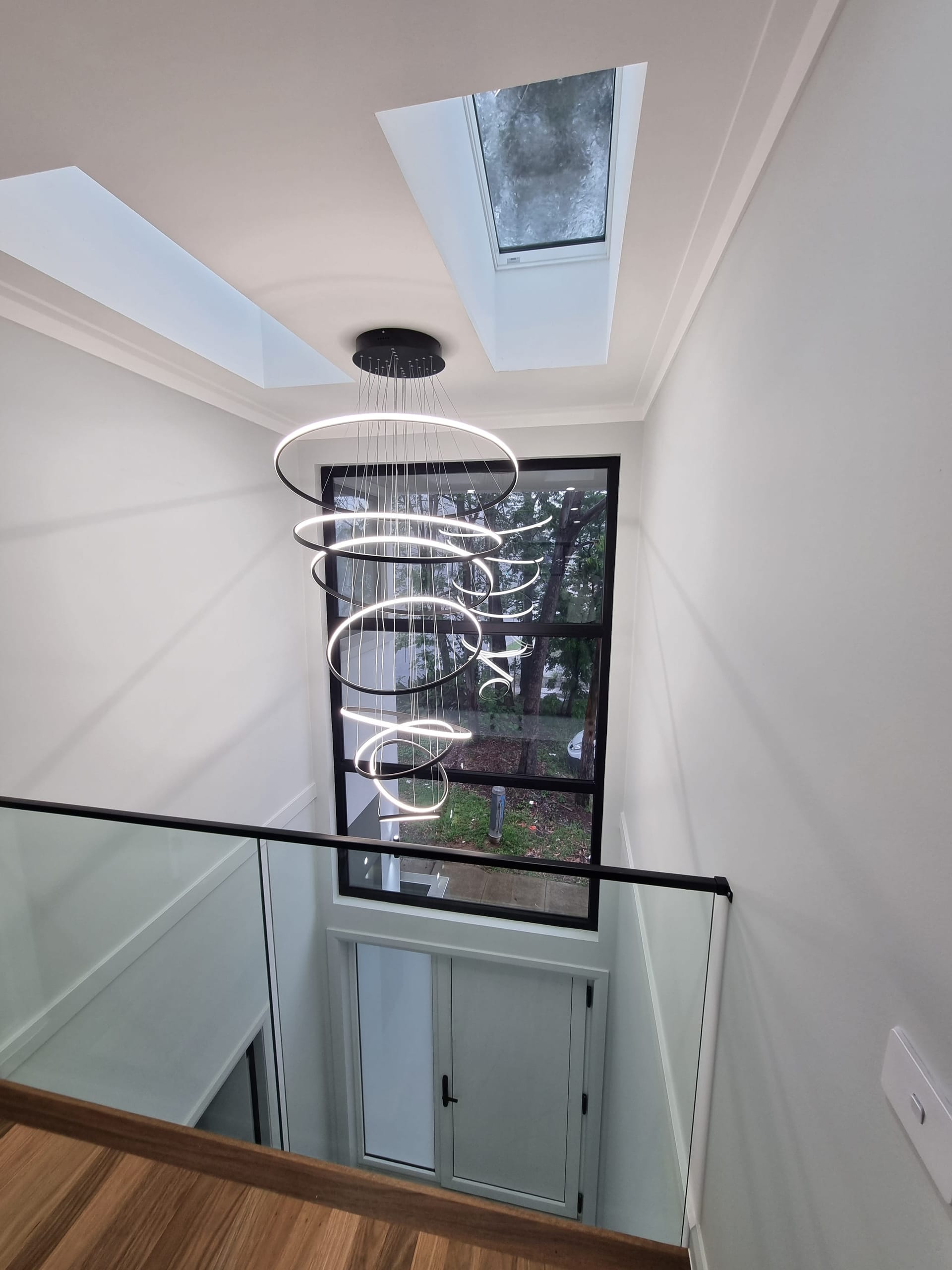 Double-height stairwell void with a multi-ring chandelier, skylights and a tall picture window to the trees