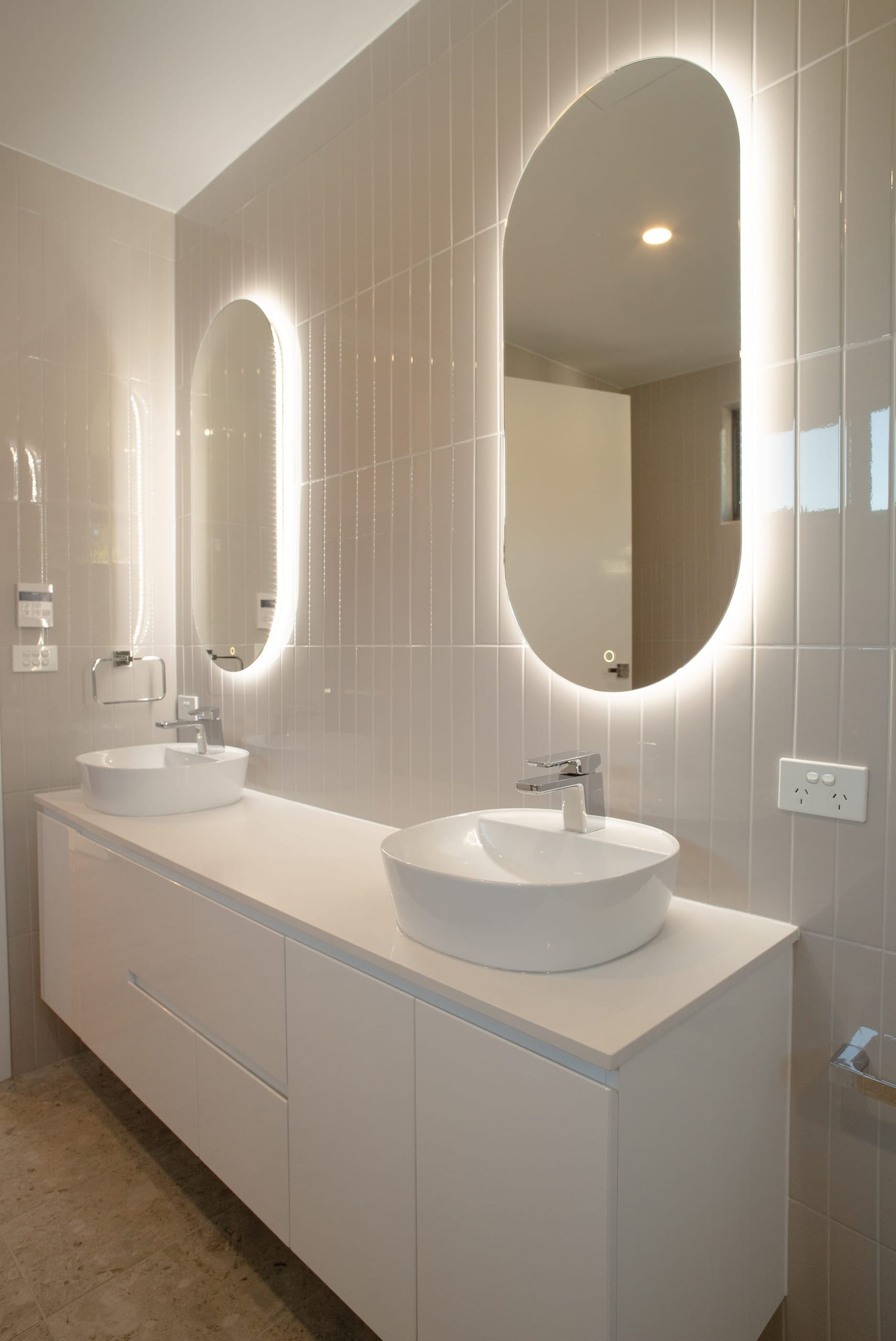 Ensuite with a double white vanity, vessel basins and oval backlit mirrors against vertical gloss tiles