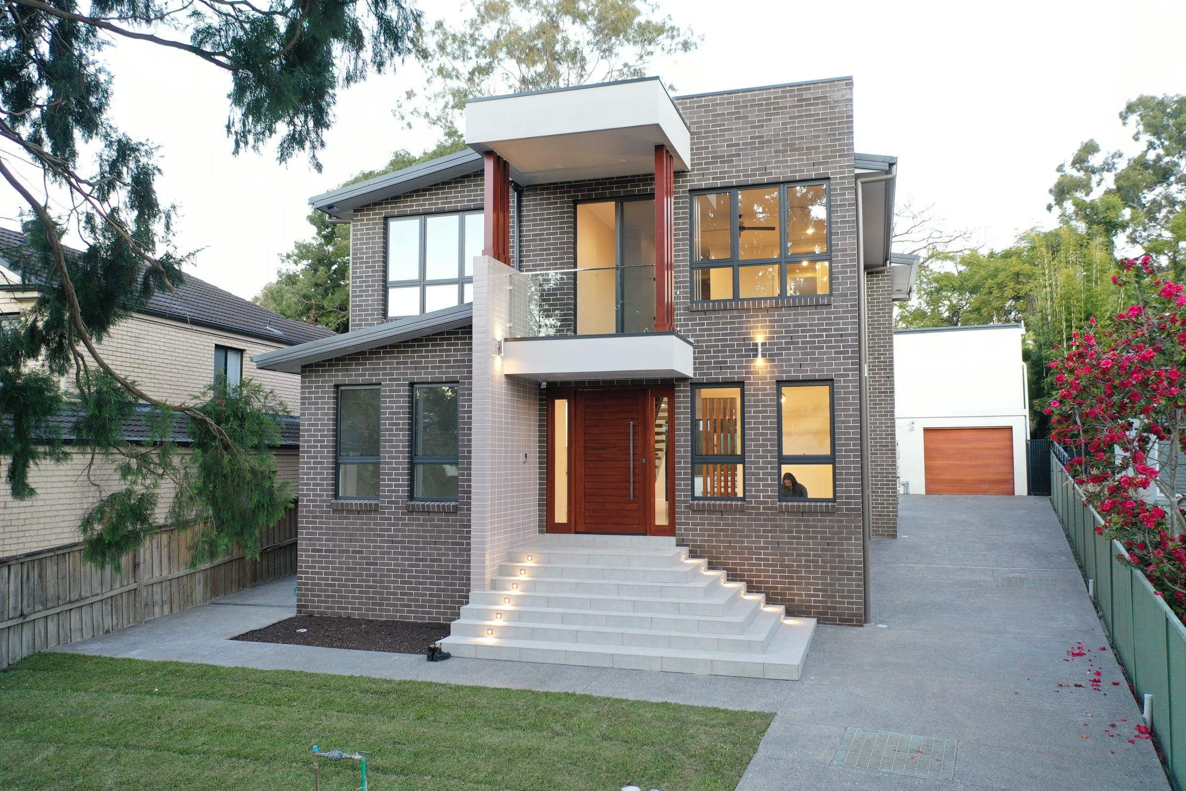 Ryde custom home at dusk, a two-storey face-brick home with a timber pivot entry, cantilevered balcony, lit entry stairs and a landscaped front lawn