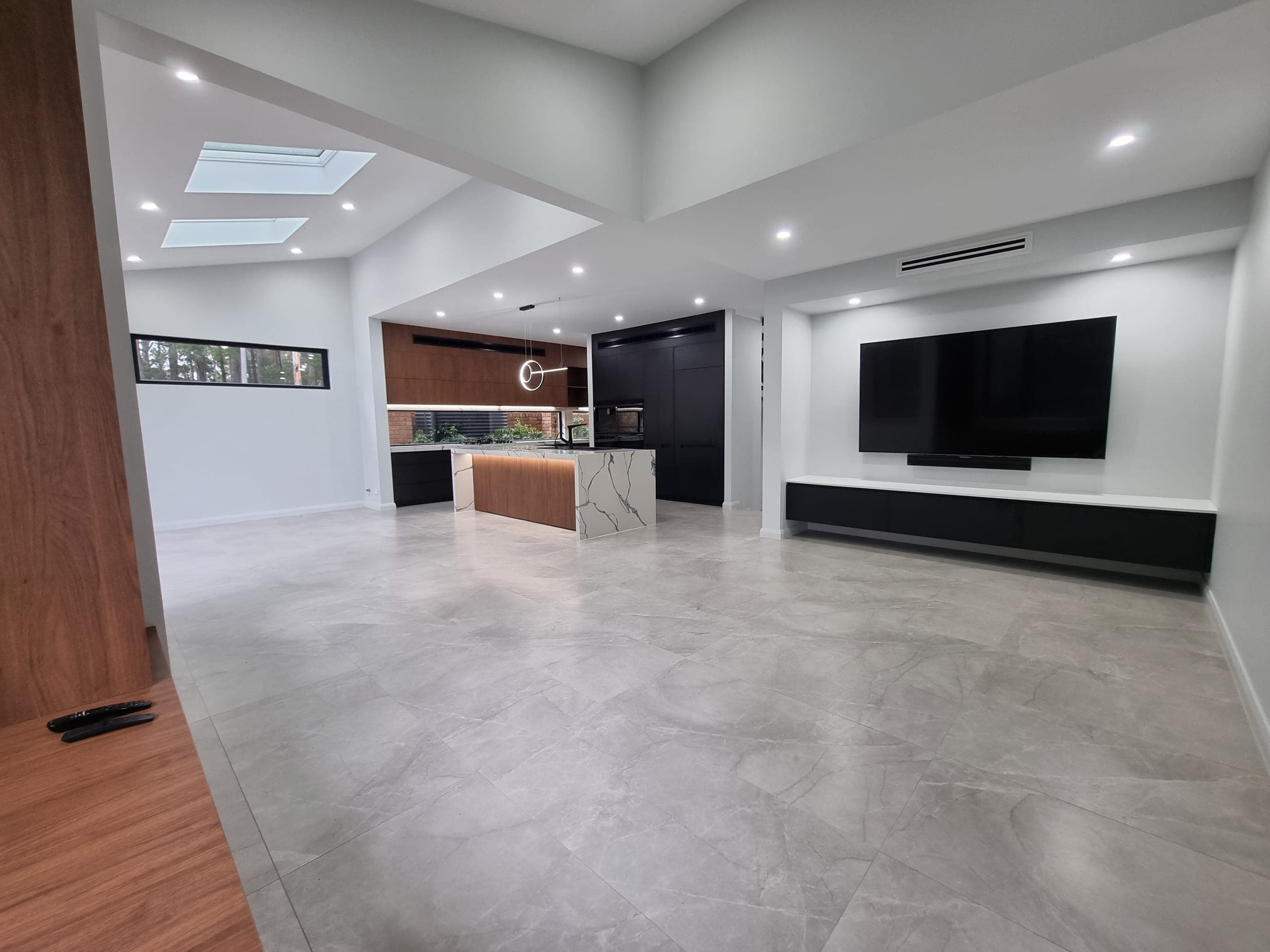 Open-plan living and kitchen with grey stone-look porcelain floors, skylights, a media wall and the kitchen beyond